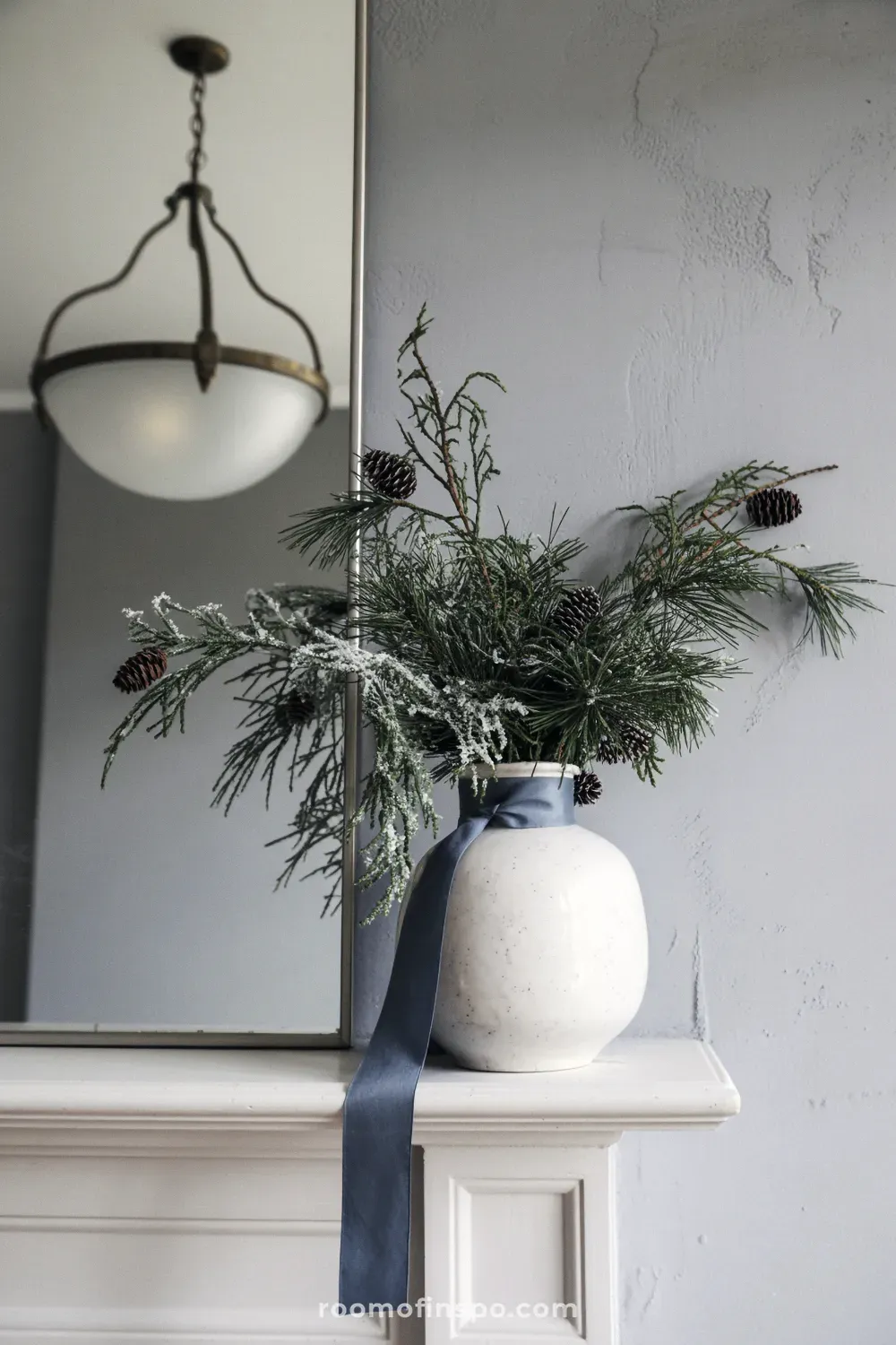 A charming winter mantel setup featuring cool blue-green foliage in a hanging glass planter above a white fireplace.
