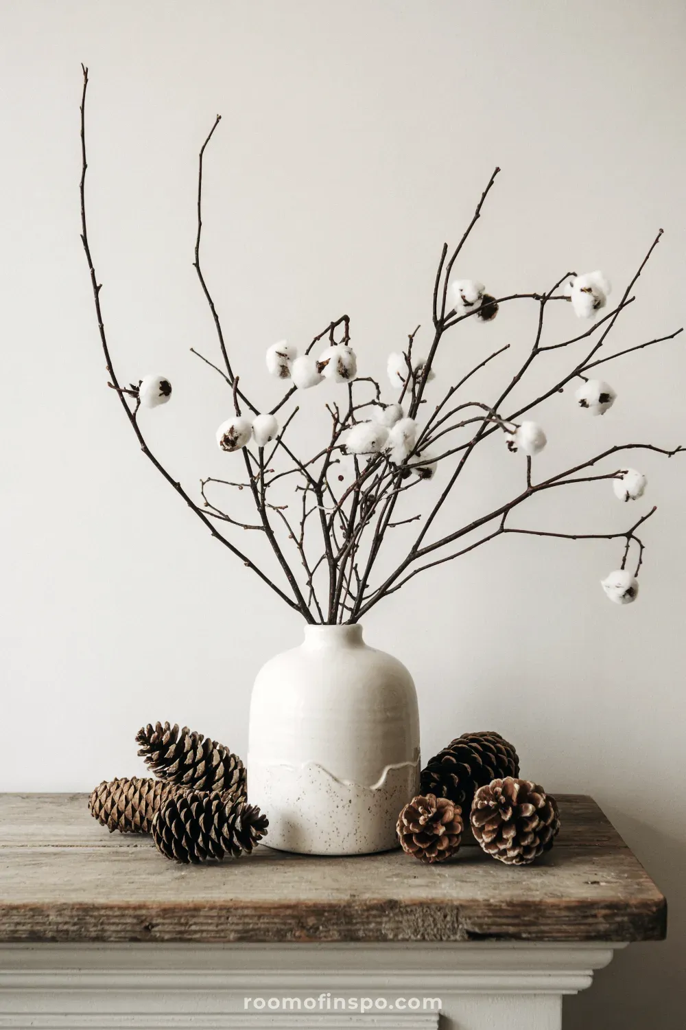Bare winter branches with pinecones are simply placed in a large white vase on a minimalist white fireplace mantel.