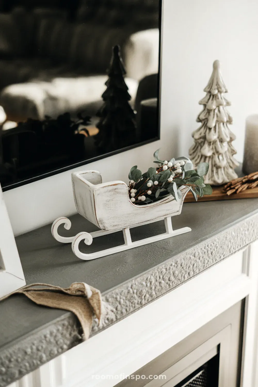 A cozy and bright winter mantel with small trees and a miniature sleigh set up by the television fireplace.