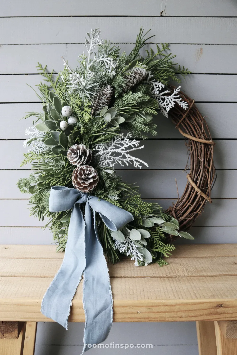 A rustic winter wreath with various green foliage, light blue ribbon, and small white berries, standing on a weathered wood bench.