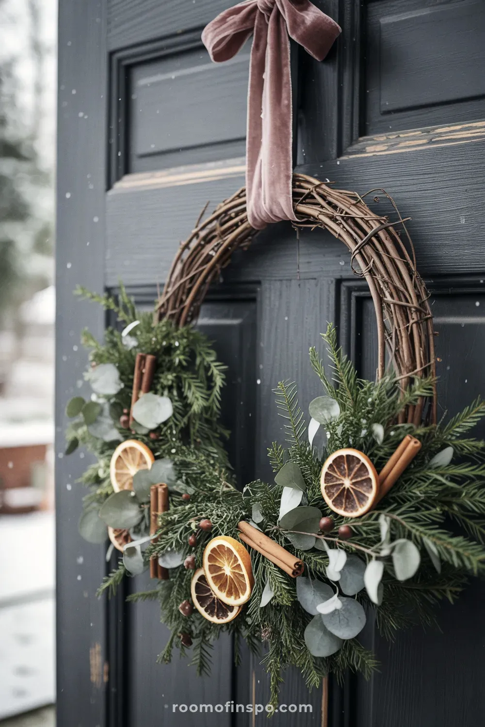 A deep green winter wreath with dried orange slices, pinecones, and red berries, hanging from a dark gray door.