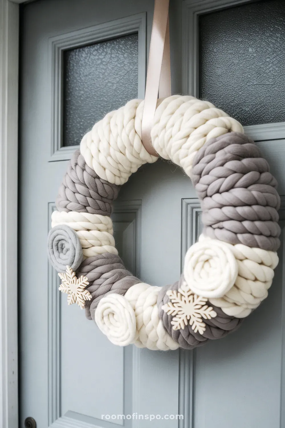 A cozy, chunky cream-colored yarn winter wreath decorated with light gray pom-poms, hanging on a pale door.