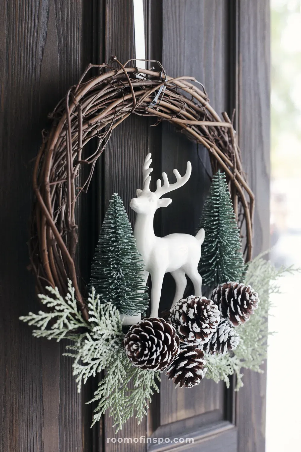 A natural twig winter wreath featuring a white deer silhouette and various snowy pine accents, hanging on a dark door.