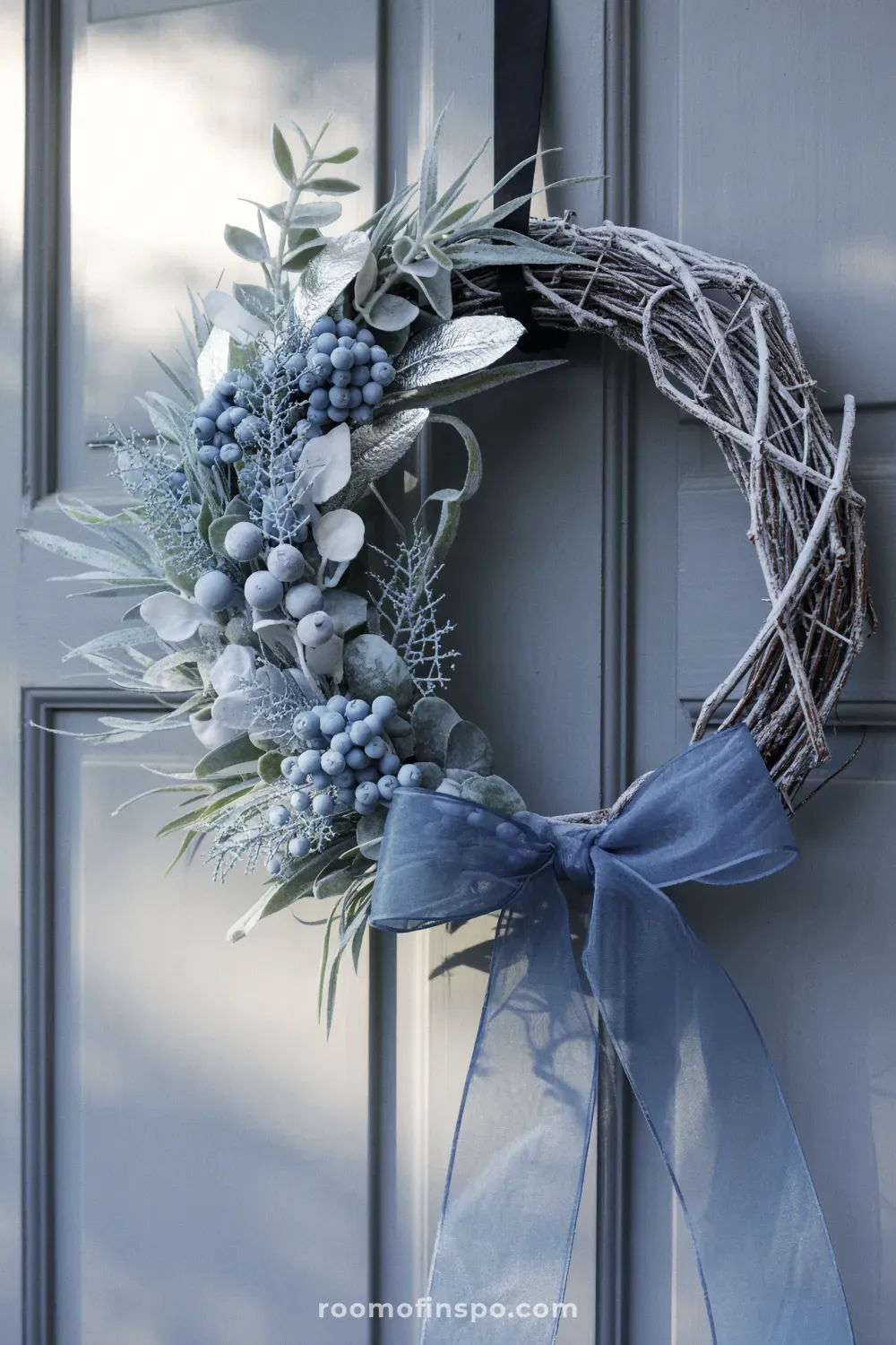 A cool-toned winter wreath with dark blue foliage, white berries, and a dark ribbon, hanging on a light blue door.