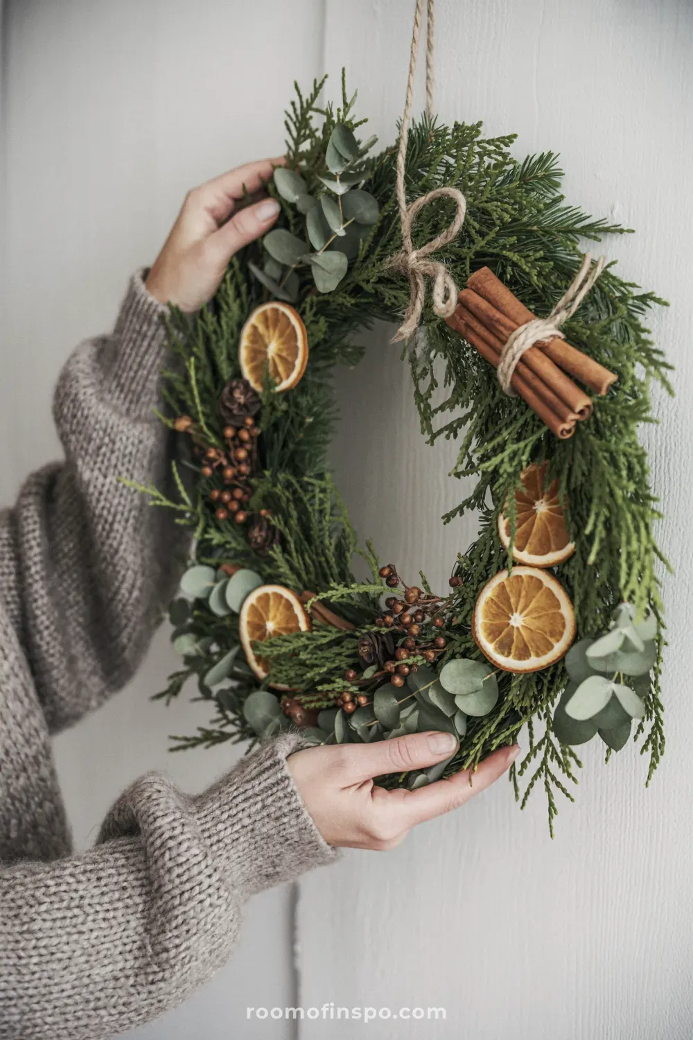 A natural winter wreath with dark green foliage, pinecones, dried orange slices, and a burlap ribbon, on a green door.