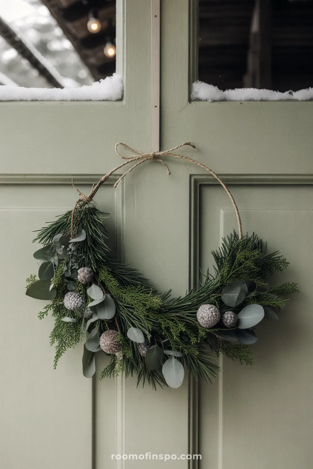 A crescent-shaped winter wreath of various green leaves and foliage, hanging on a light green panelled door.