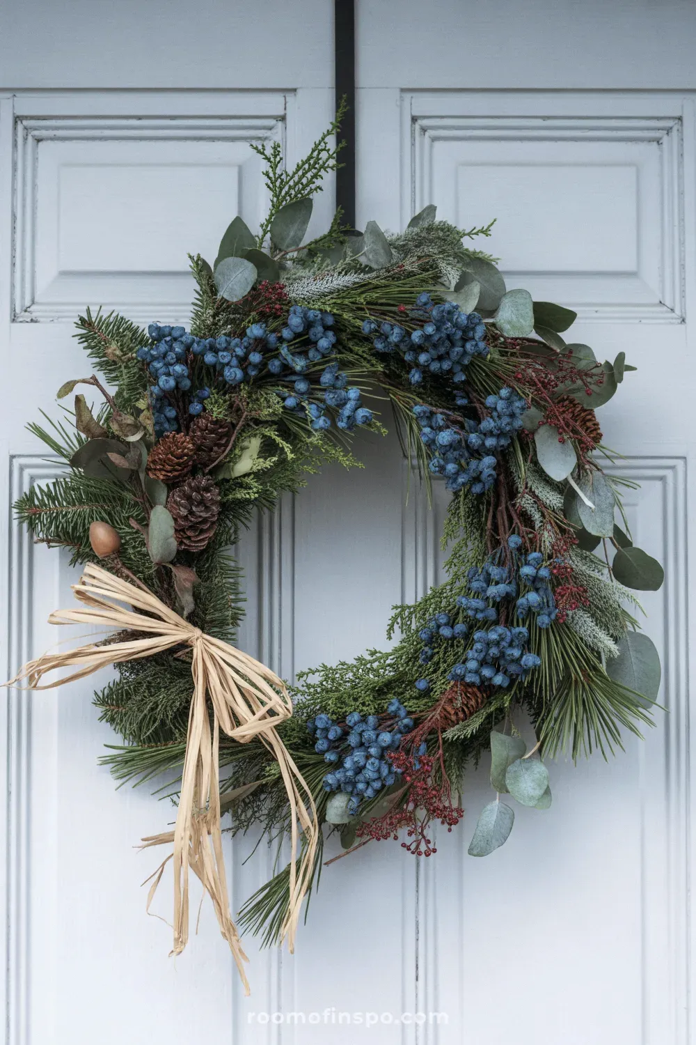 A lush winter wreath with dark green and blue-tinted foliage, accented with dark berries and small pinecones, hanging on a door.