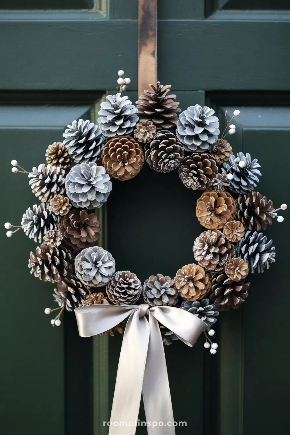 A full winter wreath covered in dark pinecones, accented with a white bow and red berries, hanging on a wooden door.