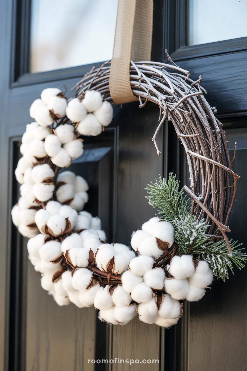 A charming cotton boll winter wreath hangs from a tan ribbon on a glossy black door, featuring frosted pine accents and twigs.