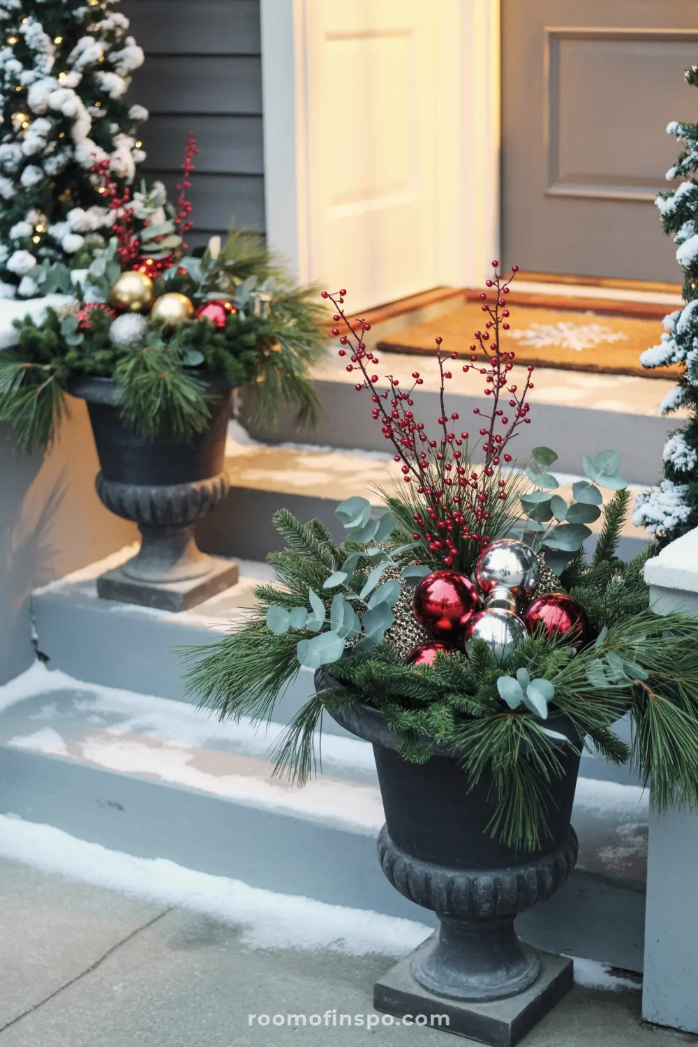 Two urns filled with festive greenery and red ornaments decorate the snow-covered front porch for Christmas.