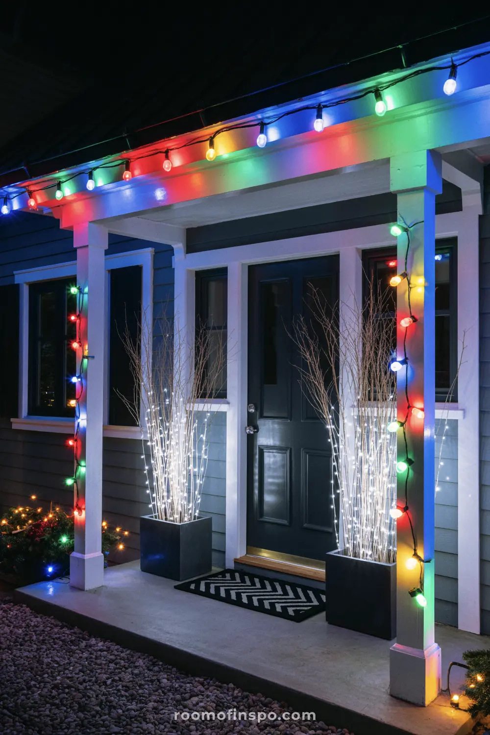 A modern front porch with colorful Christmas lights and illuminated branches in planters beside the door.