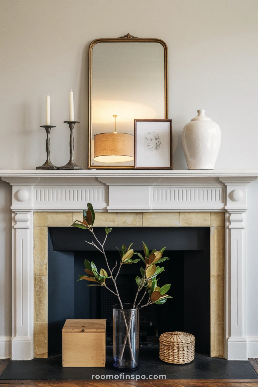 Carved white mantel with brass mirror, black candlesticks, a ginger jar, magnolia leaves, and slate hearth.