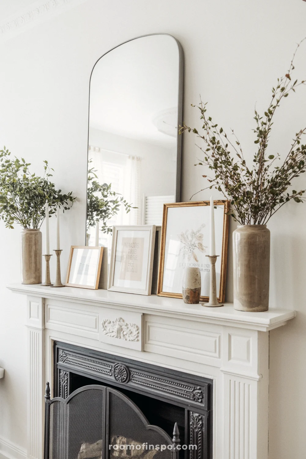Ornate white mantel with arched mirror, layered neutral frames, tall vases of dried branches, and a black screen.