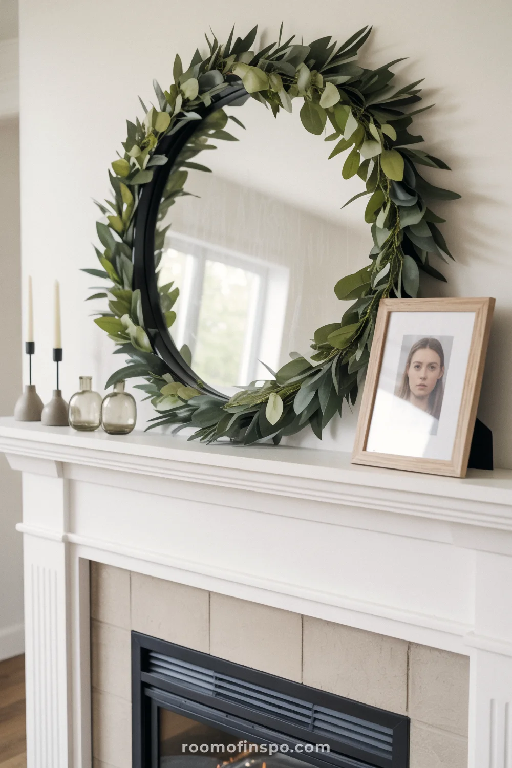 White mantel with eucalyptus garland, round black mirror, taper holders, small frames, and bud vases.