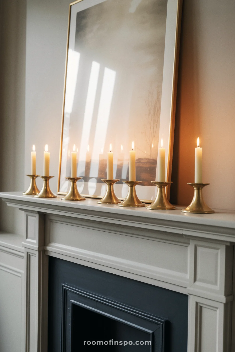 White mantel lined with brass taper holders and ivory candles, with a leaning neutral artwork behind.