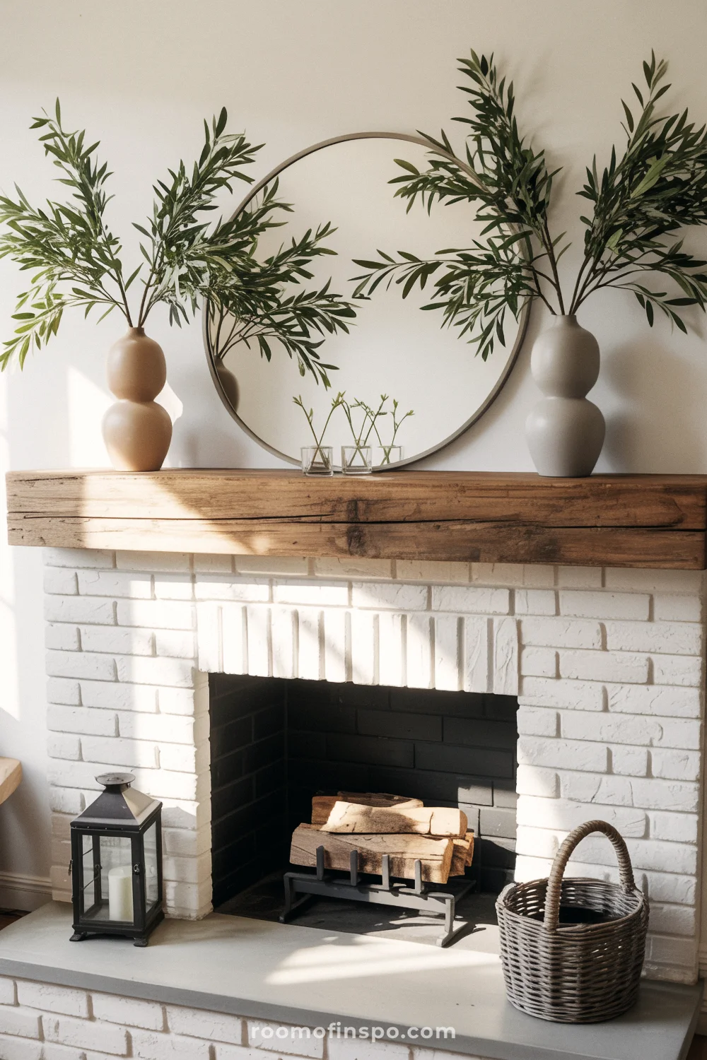 White brick fireplace with reclaimed wood mantel, two ceramic vases of olive branches, and a round black mirror.
